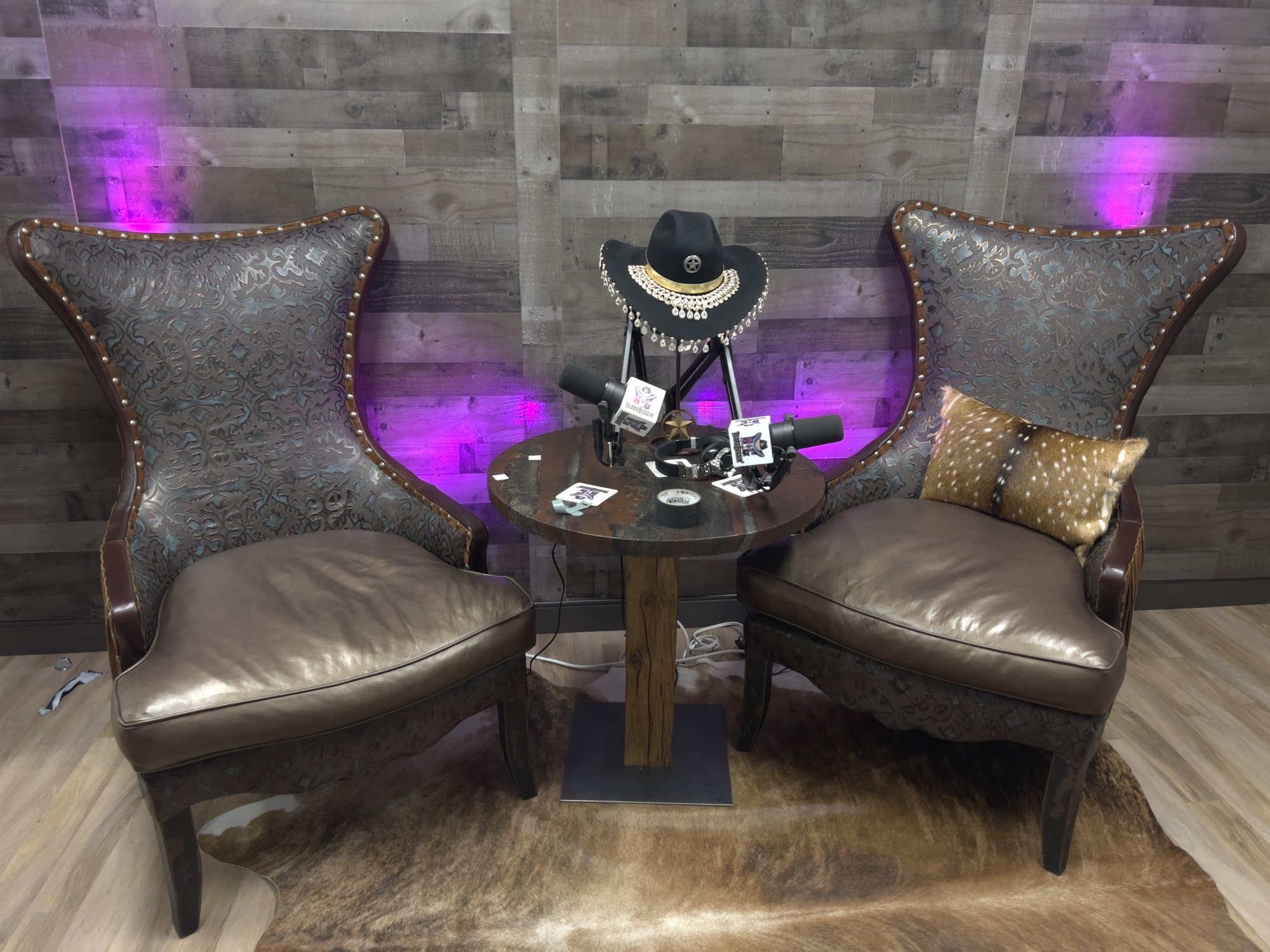 Empty podcast set with two wingback chairs facing each other under purple light, cameras ready.