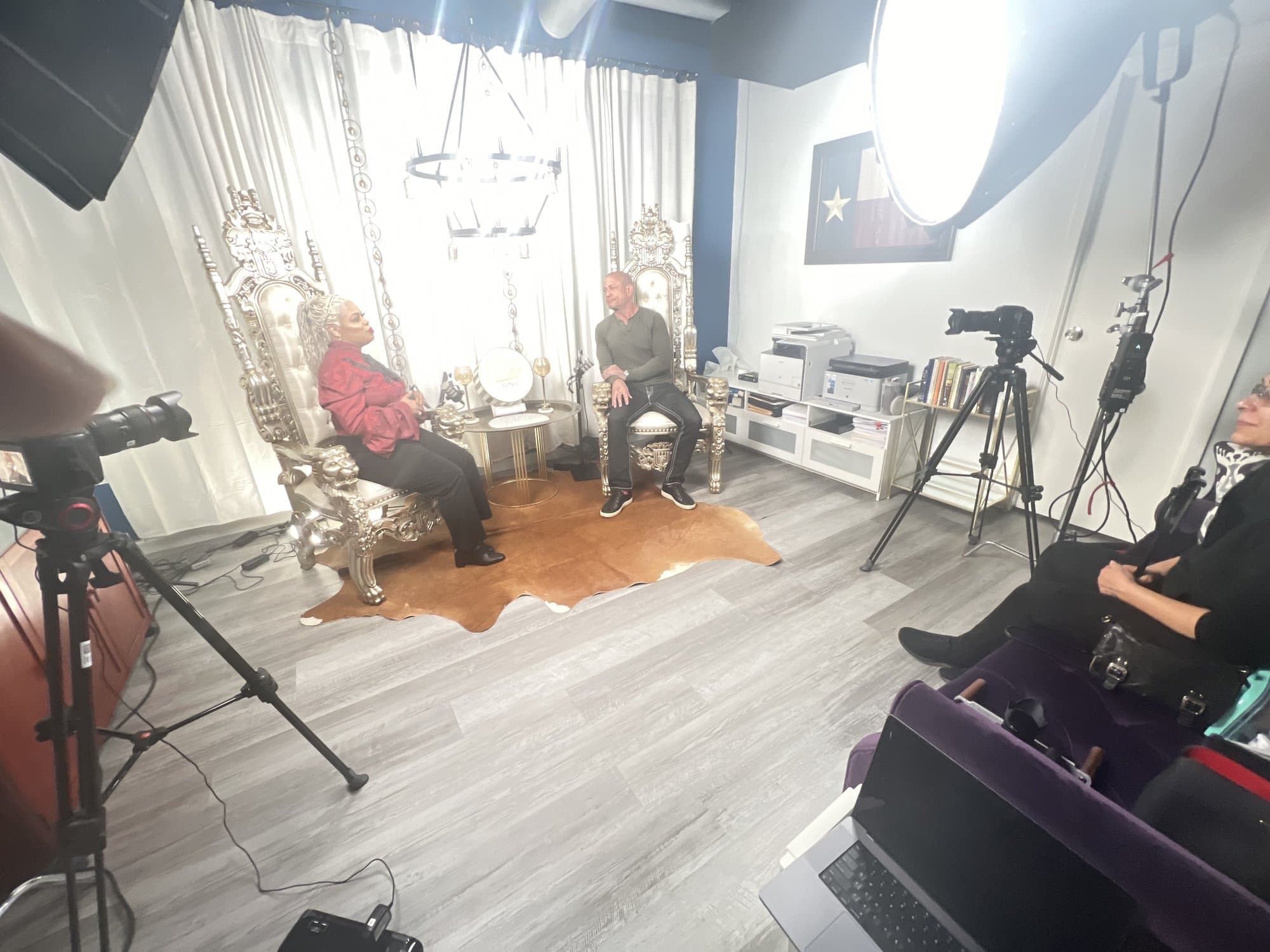 Green-wall podcast set mid-shoot, with crew, studio lights, and a clapperboard around two armchairs.